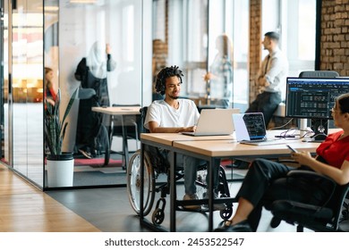 A young business group, including an African American businessman in a wheelchair, collaborates within a modern glass office, actively engaged around a computer and laptop, collectively solving - Powered by Shutterstock