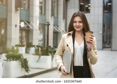 Young Business Lady Holding Laptop Under Arm Walking Down The Street Near Office Center With Cup Of Coffee Looking Busy, Eating On The Go Concept