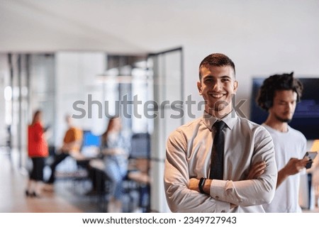 A young business leader stands with crossed arms in a modern office hallway, radiating confidence and a sense of purpose, embodying a dynamic and inspirational presence.