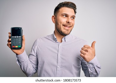Young business man with blue eyes holding dataphone payment terminal over isolated background pointing and showing with thumb up to the side with happy face smiling