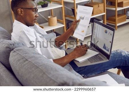 Young business man doing research. African American man sitting on comfortable couch at home, working on modern laptop computer, studying financial report, looking at digital slides, and taking notes