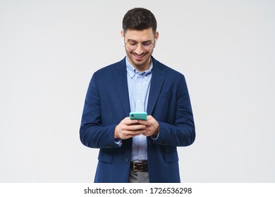 Young business man dressed in blazer and shirt, looking happily at screen of phone he is holding, reading funny messages, isolated on gray background - Powered by Shutterstock