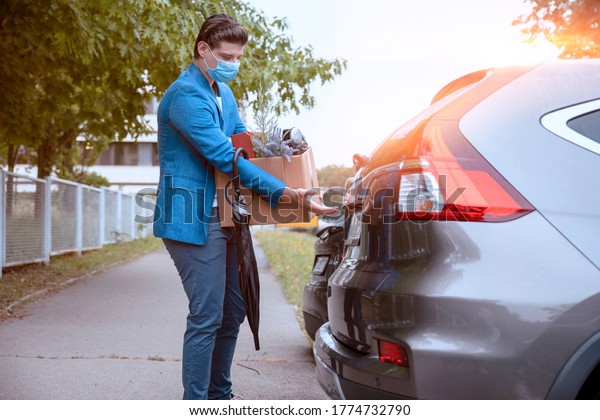 Young Business Man with face protective mask open his\
Car trunk on the Parking place, holding box of personal stuff,\
fired from the job 