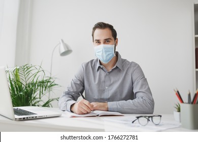 Young business man in gray shirt sterile face mask sitting at desk work on laptop pc computer in light office on white wall background. Achievement business career concept. Writing notes in notebook - Powered by Shutterstock