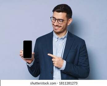 Young business man holding smartphone with blank screen, copy space included, pointing to it, advising high quality finance app, isolated on blue background