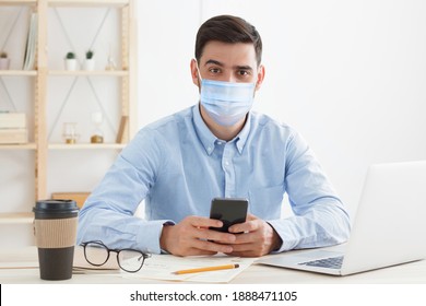 Young Business Man Sitting In Office, Wearing Mask, Practicing Social Distancing Because Covid Safety Protocol 