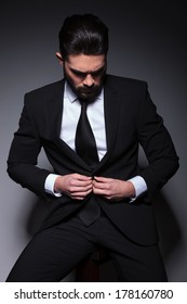 Young Business Man Sitting On A Chair And Buttoning His Suit Jacket While Looking Down, Away From The Camera. On A Dark Background