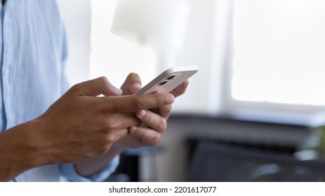 Young business man standing indoors, holding mobile phone, typing text message on mobile phone, chatting online, browsing Internet. Close up of hands. Male user, office employee making call