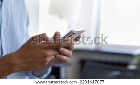 Young business man standing indoors, holding mobile phone, typing text message on mobile phone, chatting online, browsing Internet. Close up of hands. Male user, office employee making call