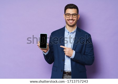 Young business man in suit, wearing eyeglasses, holding blank screen smartphone and pointing to it, copy space for advertising, isolated on purple background