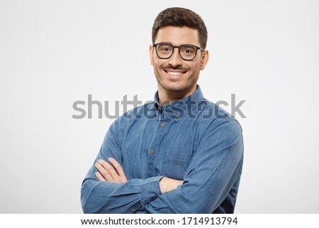 Young business man wearing blue shirt and glasses, looking at camera with positive confident smile, holding arms crossed, isolated on gray background