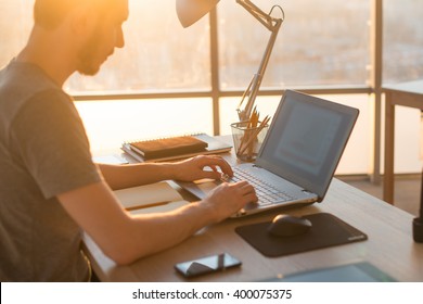 Young Business Man Working On A Laptop At His Desk In The Office, In View Of The Profile Against The Window