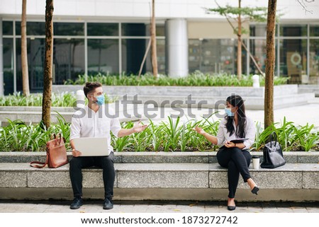 Young business people in medical masks sitting far from each other on bench outdoors when having meeting and discussing plans