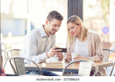 Young Business People Sitting In A Coffee Shop And Looking At Mobile Phone.