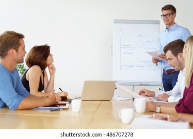 Young business team working together on a project seated around a table in the office with one man standing at a flip chart doing a presentation