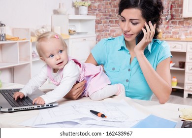 Young Business Woman With Baby In The Kitchen Working With Laptop, Speaks By Phone