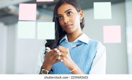 Young business woman brainstorming and planning a mind map while writing ideas on sticky notes on a glass wall in an office. Focused designer analyzing a marketing strategy and solutions for projects - Powered by Shutterstock