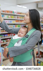 Young Business Woman Choosing Breakfast In Convenience Store While Carrying Her Baby Boy By Hipseat.