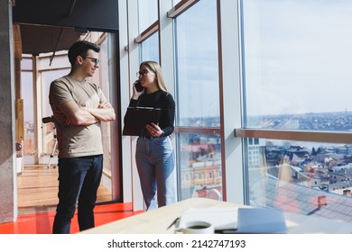 A Young Business Woman With Documents In Glasses Stands With A Male Manager While Holding A Corporate Business Meeting In A Modern Office. Business Meeting Concept. Free Space, Selective Focus