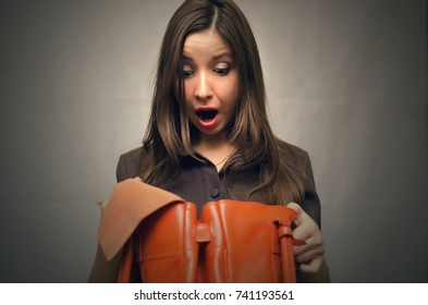 Young Business Woman In Dress With A Wide Open Mouth Looks Into The Bag She Holds In Her Hands And In Shocked From She Saw Inside. Not Enough Money To Buy. Perplexed Woman Forgot Her Wallet At Home.