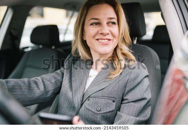 A young business woman drives a\
company car and checks her phone and looking out the\
window.