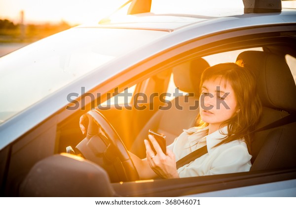 Young business woman driving car and looking\
simultaneously in her mobile\
phone