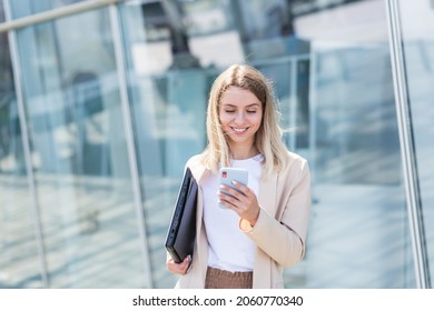 young business woman goes and uses mobile phone in her hands a urban background a modern office building. Attractive businesswoman or student Female walks the city street outdoors using a smartphone