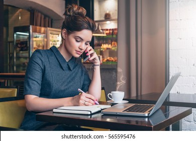 Young business woman in gray dress sitting at table in cafe, talking oncell phone while taking notes in notebook. On table laptop and cup of coffee. Student learning online. Freelancer working online. - Powered by Shutterstock