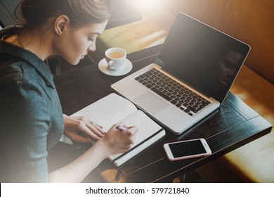 Young Business Woman In Gray Dress Sitting At Table In Cafe And Writing In Notebook. On Table Is Laptop, Smartphone And Cup Of Coffee. Freelancer Working In Coffee Shop. Student Learning Online.