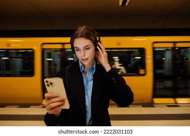 Young Business Woman In Headphones Holding Mobile Phone While Waiting For Train At Subway Station