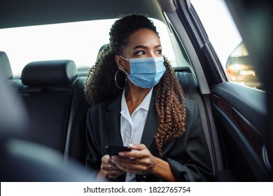 Young Business Woman In A Mask Checking Her Mobile Cell Phone On A Backseat Of A Taxi During Covid-19 Quarantine. Business Trips During Pandemic, New Normal And Coronavirus Travel Safety Concept.