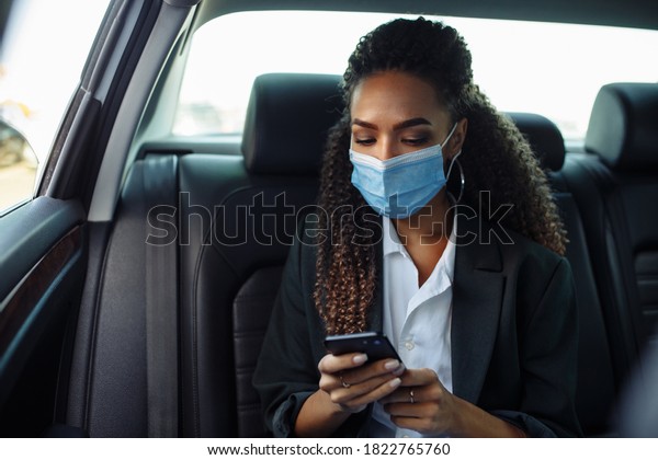 Young business woman in a mask checking her\
mobile cell phone on a backseat of a taxi during covid-19\
quarantine. Business trips during pandemic, new normal and\
coronavirus travel safety\
concept.