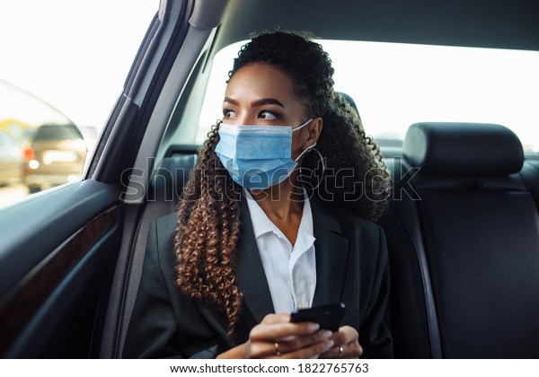 Young business woman in a mask checking her
mobile cell phone on a backseat of a taxi during covid-19
quarantine. Business trips during pandemic, new normal and
coronavirus travel safety
concept.
