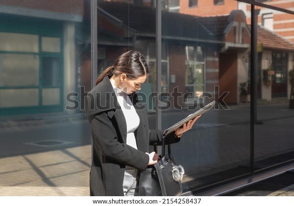 Young business woman on formal wear standing\
outdoor in front of the building searching her office keys or car\
keys in her purse that she lost. Person search for her smartphone\
in her handbag.