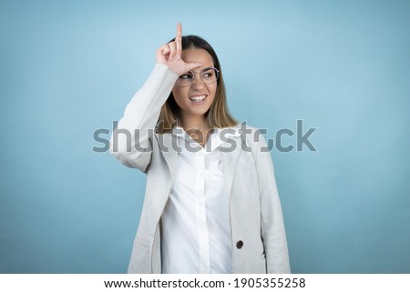 Young business woman over isolated blue background making fun of people with fingers on forehead doing loser gesture mocking and insulting.