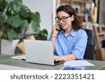 Young business woman professional employee, entrepreneur sitting at desk working online on laptop making call on cell phone taking to client regarding online website sales order or marketing work.