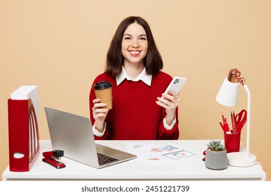 Young business woman in red sweater sit work at office desk with pc laptop hold takeaway delivery cup coffee to go use mobile cell phone isolated on plain beige background. Achievement career concept