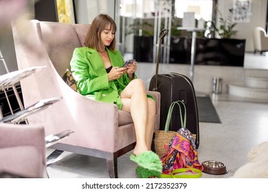 Young Business Woman Sits In Comfortable Sofa And Uses Phone In Lobby While Waiting For Check In At Luxury Hotel. Business Trip And Style Concept