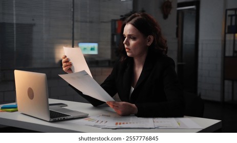 Young business woman sits at desk works late night in office, using laptop, reading documents, concentrated focused expression, side shot. - Powered by Shutterstock