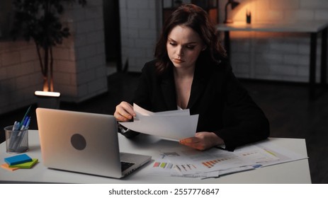 Young business woman sits at desk works late night in office, using laptop, reading documents, serious focused expression, middle shot. - Powered by Shutterstock