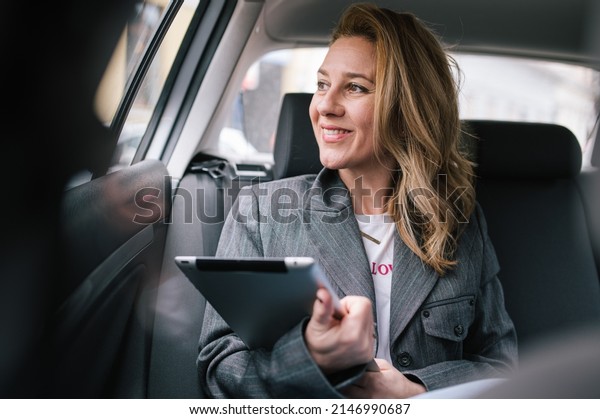 A young business woman\
sitting in the back seat of a car uses a tablet and looks out the\
window.