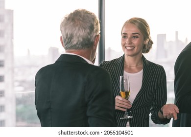 Young Business Woman Talking To Her Older Business Partner While Holding A Champagne Glass In Her Hand. She Is Happy Smiling. The Scene Behind Is The City Scape.