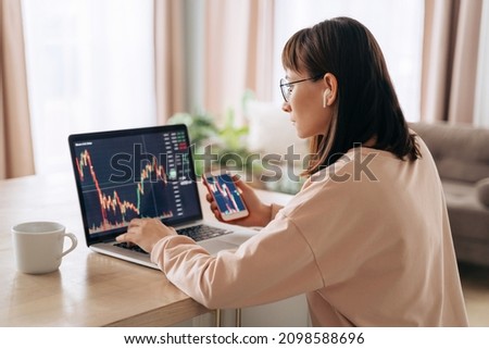 Young business woman trader analyst looking at laptop monitor, holding smartphone. Investor broker analyzing indexes, trading online investment data on cryptocurrency stock market graph at home