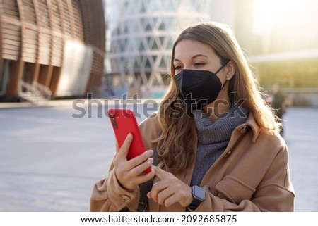 Young business woman wearing black medical face mask using mobile phone app in city street
