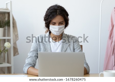 Young business woman wearing face mask working on computer seated at workplace desk in office room protecting herself from getting grippe vs COVID-19 corona virus pandemic infectious disease concept