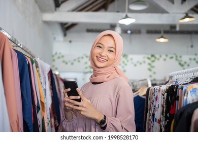 A Young Business Woman Wearing A Hijab Holding A Cellphone Is Standing In A Boutique Shop