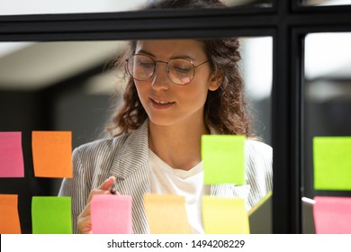 Young business woman write tasks creative ideas on sticky notes on glass board, female corporate leader planning project on post it sticky notes organize work on stickers on scrum glass window wall - Powered by Shutterstock