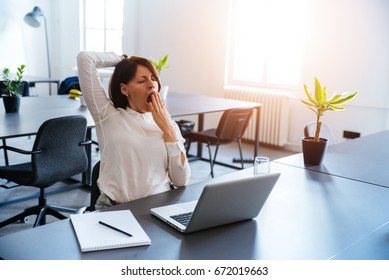 Young business woman yawning at a modern office desk in front of laptop