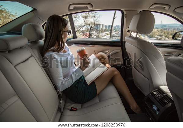 A young business women drives on\
back seats, drinking a coffee and looking out the window, with note\
in the lap. She looks so attractive in skirt.\

