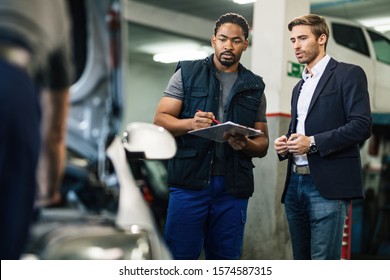Young businessman and African American car mechanic talking while examining car at auto repair shop.  - Powered by Shutterstock